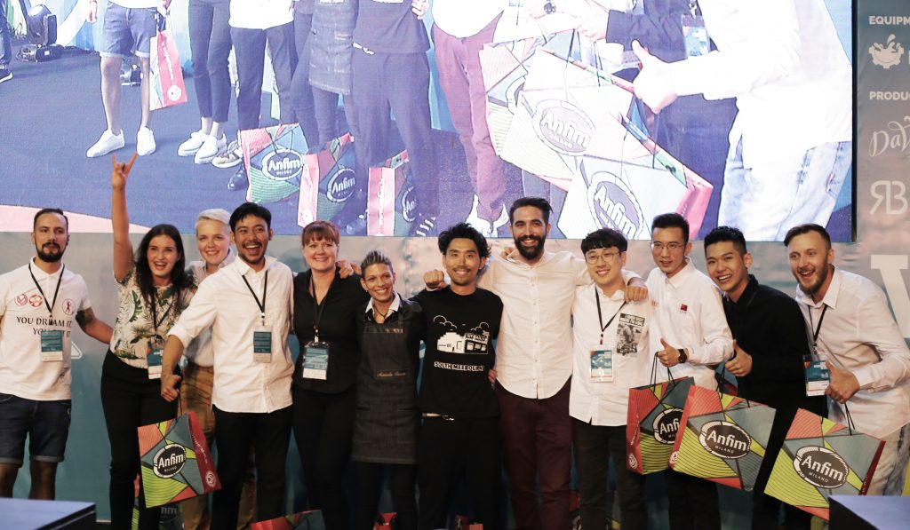 Twelve semi-finalists for the World Latte Art Championship stand in front of a large screen & stage wall.